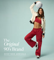 A woman wearing a cream zip-up jacket, red sports bra, red wide-leg track pants, and sneakers poses against a light blue background while balancing a white basketball on her head. She stands on one leg with a playful expression. Text: The Original 90's Brand. New Arrivals - Shop Now