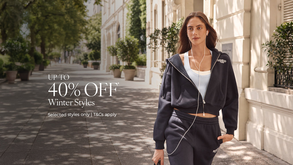 A woman wearing a navy Lorna Jane cropped zip hoodie, light blue sports tank, and matching track pants while walking outdoors with wired earphones. Text: Up To 40% Off* Winter Styles. Selected Styles Only | T&Cs apply - Shop Now