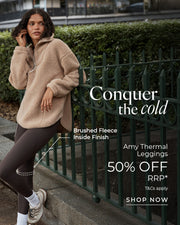 Woman wearing a beige brushed fleece half-zip pullover and dark brown thermal leggings, leaning against a green fence in an urban park. Brushed Fleece Inside Finish. Text: Conquer The Cold. Amy Thermal Leggings 50% Off RRP* T&Cs apply - Shop Now
