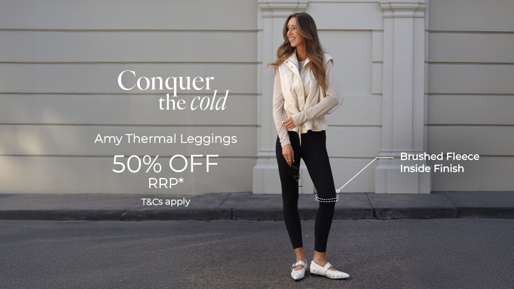A woman wearing a white quilted vest, cream long-sleeve top, black Amy Thermal Leggings, and white flats, standing on a city street. Text: Conquer The Cold. Amy Thermal Leggings 50% Off RRP* Brushed Fleece Inside Finish. T&Cs Apply - Shop Now