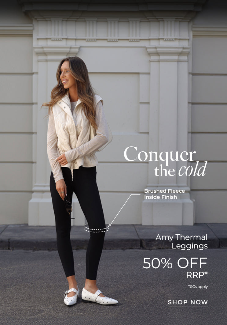A woman wearing a white quilted vest, cream long-sleeve top, black Amy Thermal Leggings, and white flats, standing on a city street. Text: Conquer The Cold. Amy Thermal Leggings 50% Off RRP* Brushed Fleece Inside Finish. T&Cs Apply - Shop Now