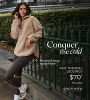 Woman wearing a beige brushed fleece half-zip pullover and dark brown thermal leggings, leaning against a green fence in an urban park. Brushed Fleece Lining. $70* T&Cs apply. Shop Now. 