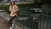 Woman wearing a beige brushed fleece half-zip pullover and dark brown thermal leggings, leaning against a green fence in an urban park. Brushed Fleece Lining. $70* T&Cs apply. Shop Now.