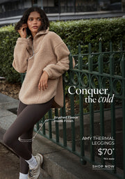 Woman wearing a beige brushed fleece half-zip pullover and dark brown thermal leggings, leaning against a green fence in an urban park. Brushed Fleece Lining. $70* T&Cs apply. Shop Now. 