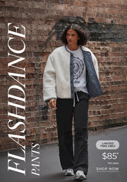 A woman wearing black Flashdance pants, a grey graphic sweatshirt, and a cream Lorna Jane everyday teddy quarter zip sweat while walking along a brick-lined street. Text: Flashdance Pants Limited Time Only $85* T&Cs apply - Shop Now