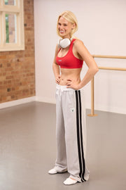 Blonde woman in a red sports bra and grey striped track pants with headphones around her neck, smiling in a dance studio.