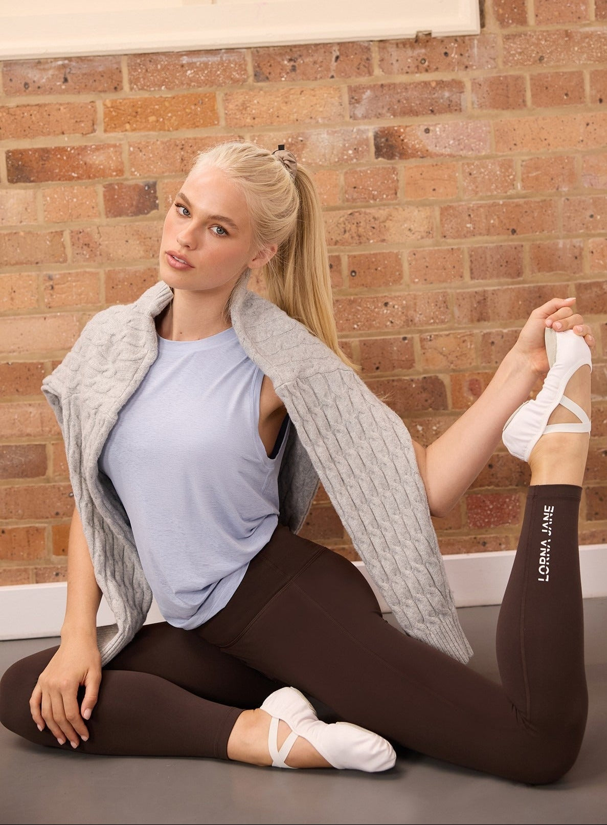 Blonde woman in a light blue tank top and brown leggings with a grey cable knit cardigan draped over her shoulders, seated stretching against a brick wall.