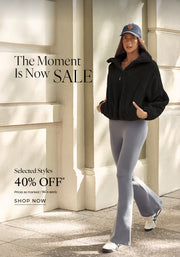 A woman wearing a black fleece jacket, gray flared leggings, white sneakers, and a blue cap while walking outdoors. Text: The Moment Is Now Sale. Selected Styles 40% Off* Prices as marked | T&Cs Apply - Shop Now