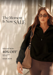 A woman walking outdoors along a sunlit city walkway, wearing a black half-zip teddy fleece pullover, brown leggings, sunglasses, and carrying a shoulder bag. Text: The Moment Is Now SALE. Selected Styles 40% OFF* Prices as marked - T&Cs apply. Shop Now