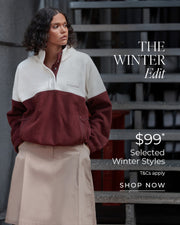 Woman with curly hair wearing a cream and burgundy color-block fleece pullover with a burgundy collared shirt underneath and a beige pleated skirt, standing on an industrial staircase. Text overlay reads 'The Winter Edit — $110 Selected Winter Styles, T&Cs apply, Shop Now.'