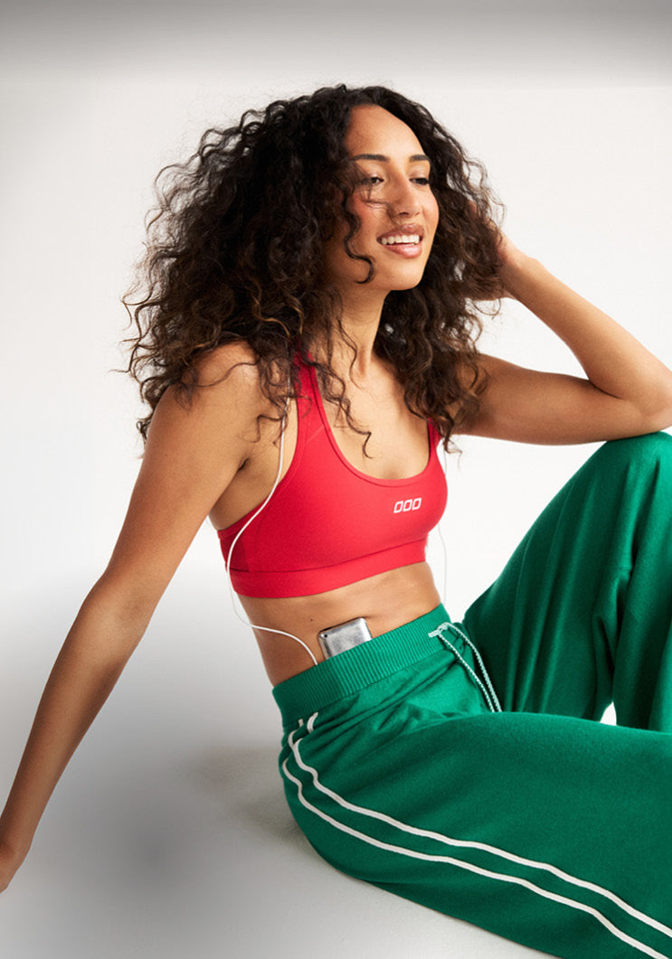 Woman wearing a red Lorna Jane sports bra and green track pants, seated against a grey studio backdrop. Text: Just Landed: New Arrivals. Shop The Collection.