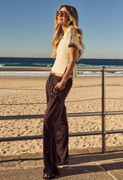 Woman standing on a beachside promenade wearing a cream short-sleeve knit top with contrast trim and dark wide-leg track pants with snap-button sides, with the ocean in the background.