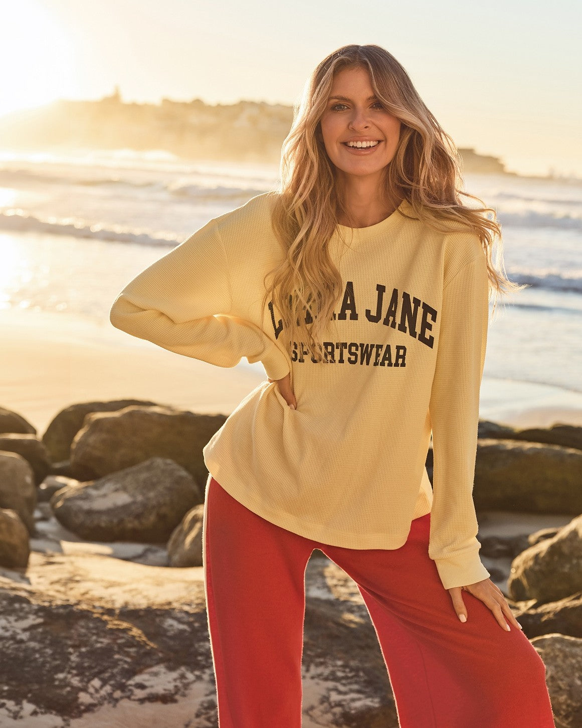 Woman standing by the beach wearing a cream graphic sweatshirt with red wide-leg pants, smiling with the ocean in the background.