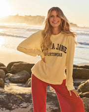 Woman standing by the beach wearing a cream graphic sweatshirt with red wide-leg pants, smiling with the ocean in the background.