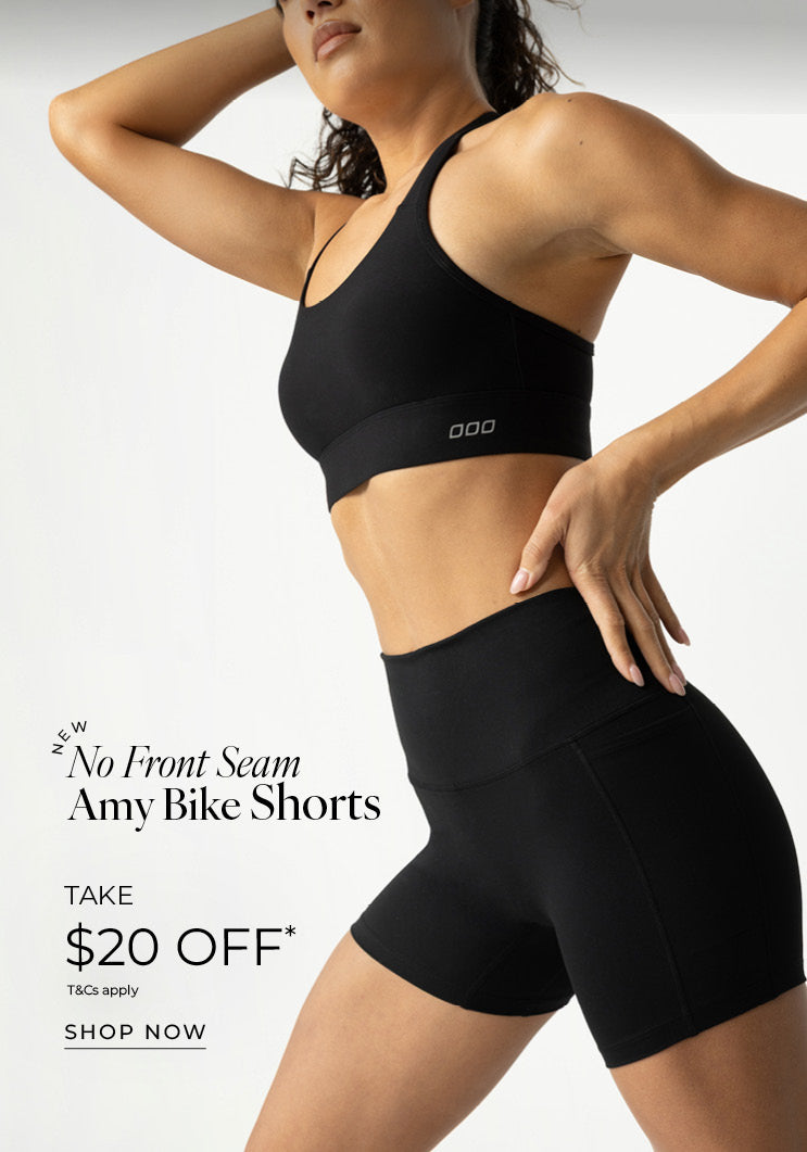 A woman posed against a plain grey studio backdrop, cropped from the chin down, wearing an all-black Lorna Jane set, a strappy sports bra and high-waisted bike shorts with her one hand on her hip, the other raised behind her head. Text: New No Front Seam Amy Bike Shorts Take $20 Off* T&Cs apply - Shop now