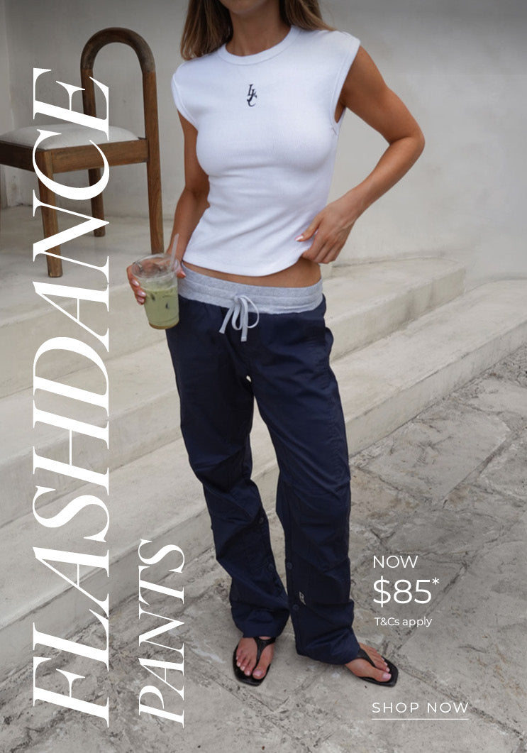 Woman wears navy Flashdance Pants with a grey drawstring waistband, styled with a white sleeveless top, sunglasses, and a navy cap, holding an iced drink in a bright, sunlit stone setting. Text: Flashdance Pants Now $85* T&Cs apply - Shop Now