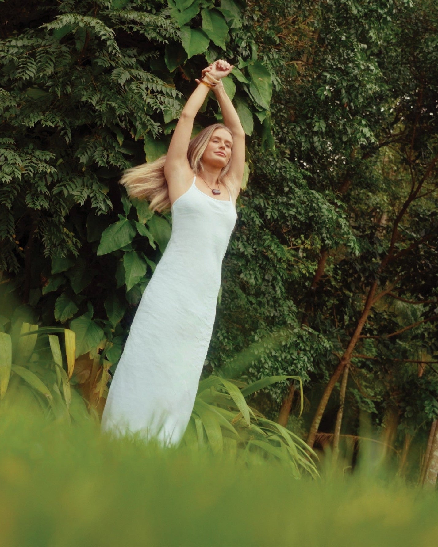 A blonde woman stretches both arms overhead in a long white linen slip dress, standing among tall grass and dense green tropical trees.