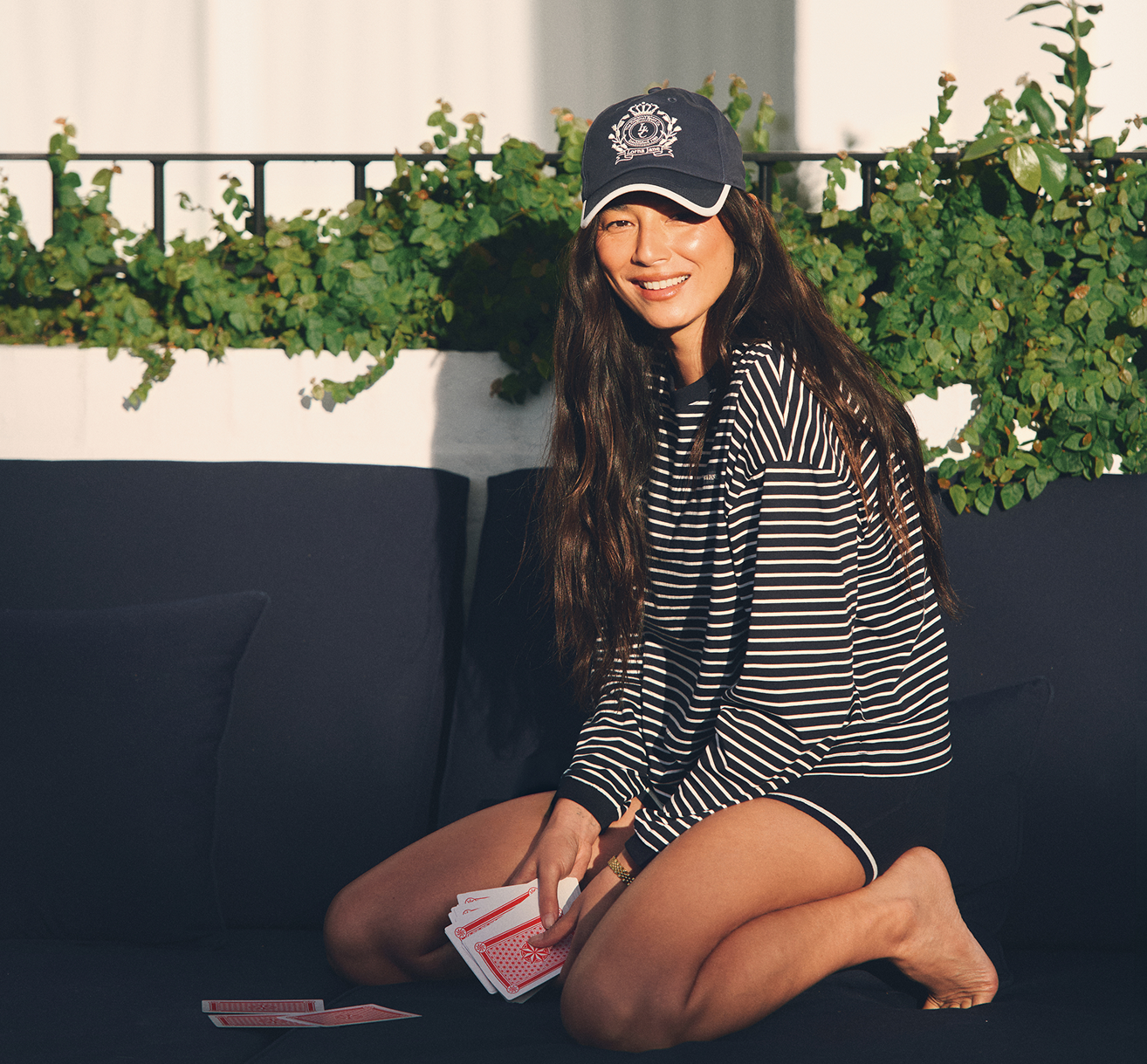 A smiling dark-haired woman sits on a navy outdoor sofa holding a hand of playing cards, wearing a navy-and-white striped long-sleeve top and a navy Lorna Jane crest cap, with greenery behind her.