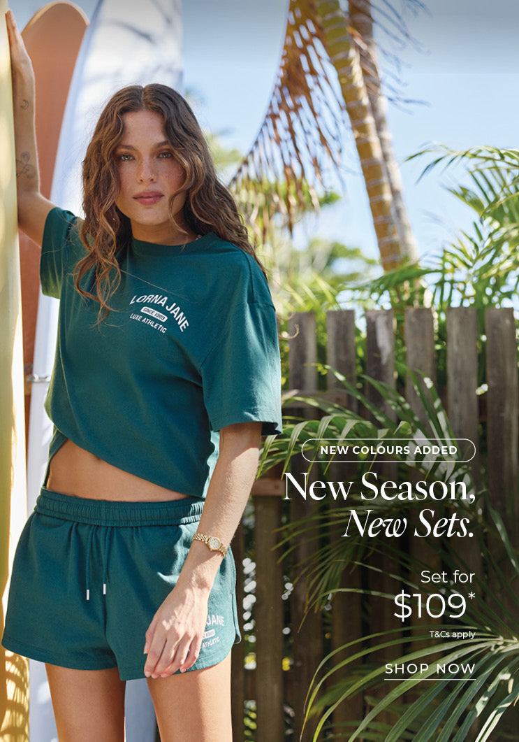 Woman in a deep teal Lorna Jane cropped tee and matching shorts, standing beside surfboards in a tropical garden. Text: New Colours Added. New Season, New Sets. Set for $109* T&Cs apply - Shop Now