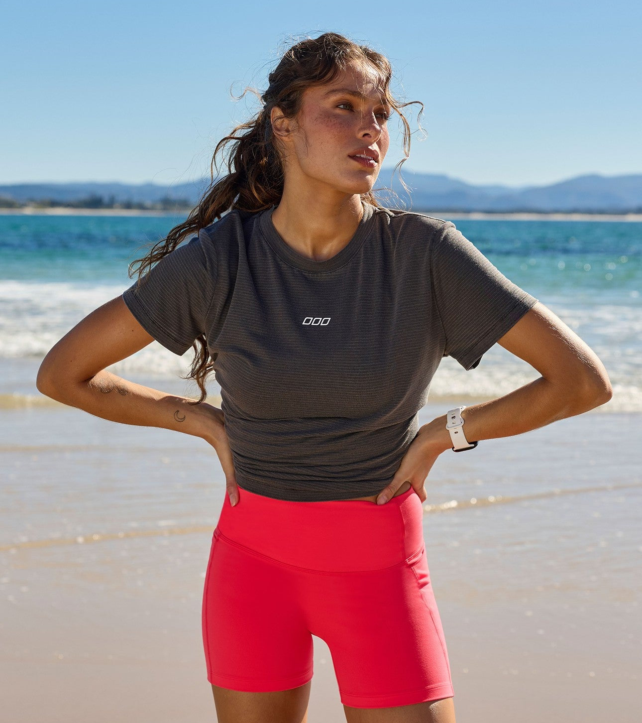 Woman in a charcoal ribbed LJ logo tee tucked into coral-red high-waisted bike shorts, hands on hips on the beach.