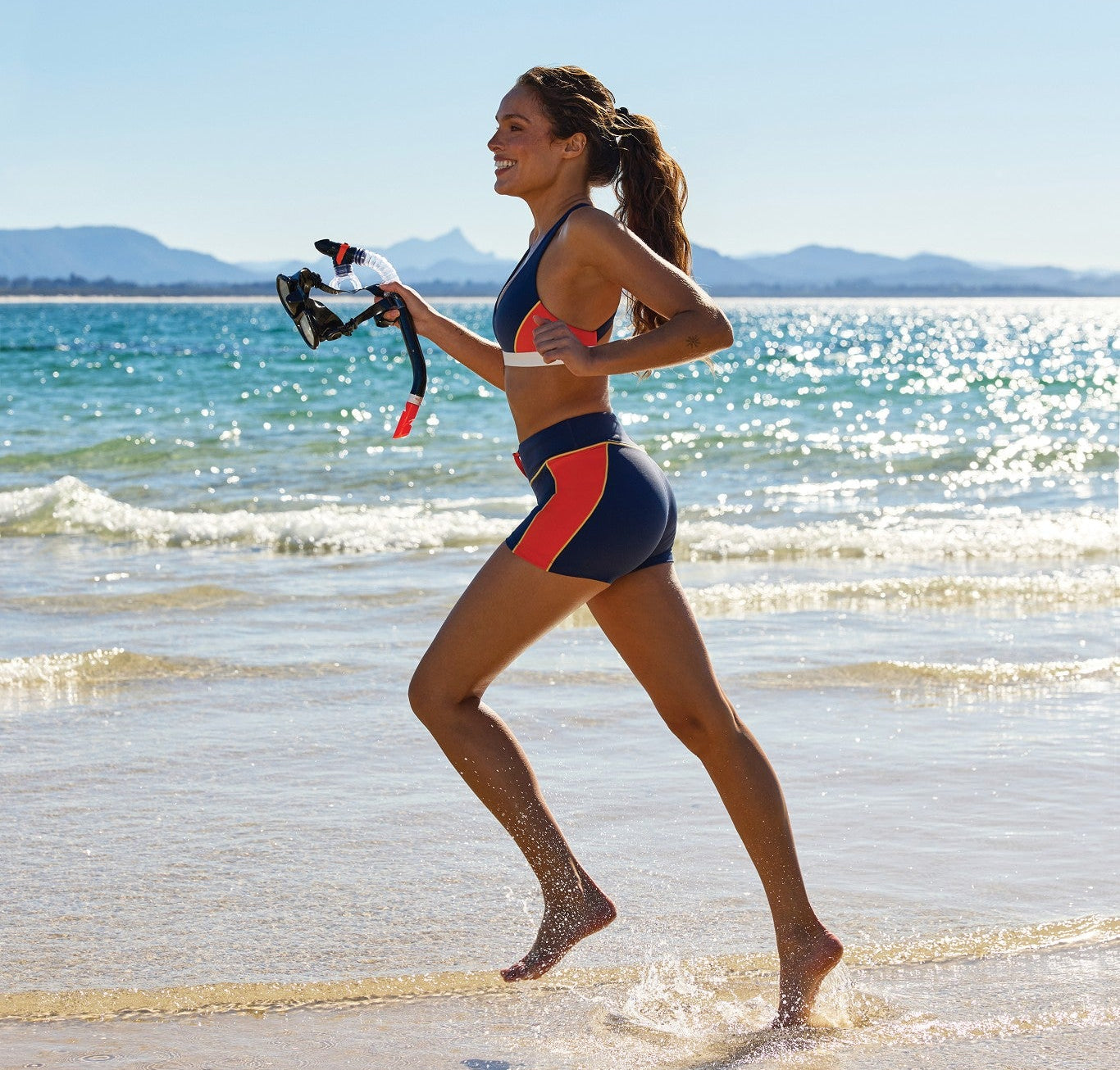 Woman running along the shoreline in a navy, red and white colour-block sports bra and bike shorts, holding a snorkel and mask.