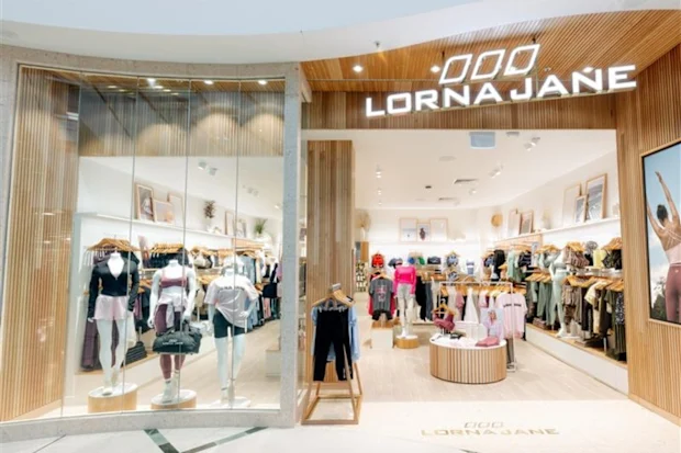 Front view of a Lorna Jane retail store in a shopping mall, featuring a modern wood-paneled entrance, large glass display windows with activewear mannequins, and a bright, spacious interior showcasing women's athletic apparel and accessories on clothing racks and display tables.