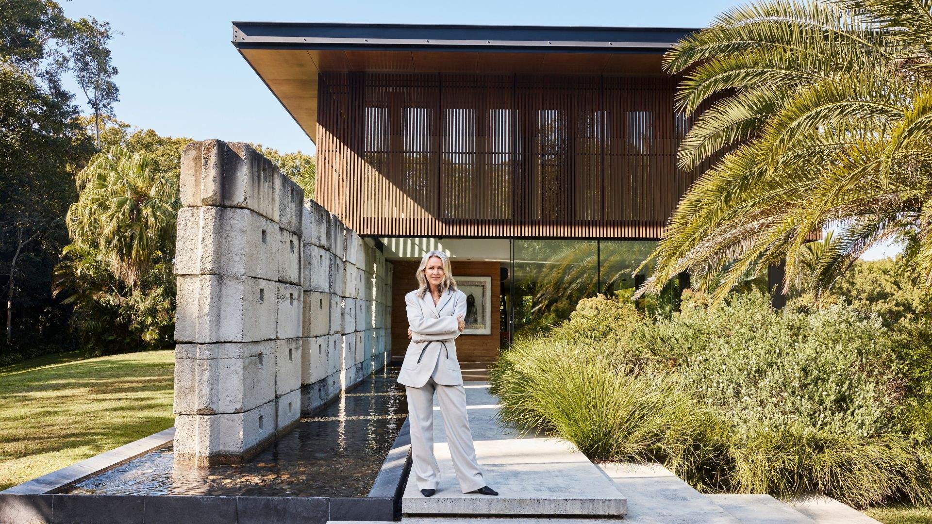 Lorna Jane in a light-colored pantsuit stands with her arms crossed outside a modern luxury retreat, surrounded by landscaped gardens, a reflecting pool, and contemporary architecture.