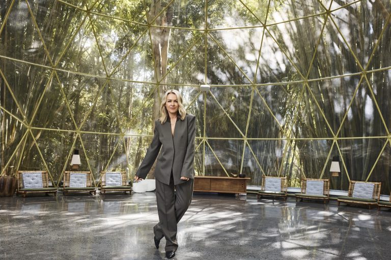 Lorna Jane in a tailored charcoal pantsuit inside a spacious glass-domed pavilion surrounded by lush bamboo, with lounge seating arranged around the perimeter.