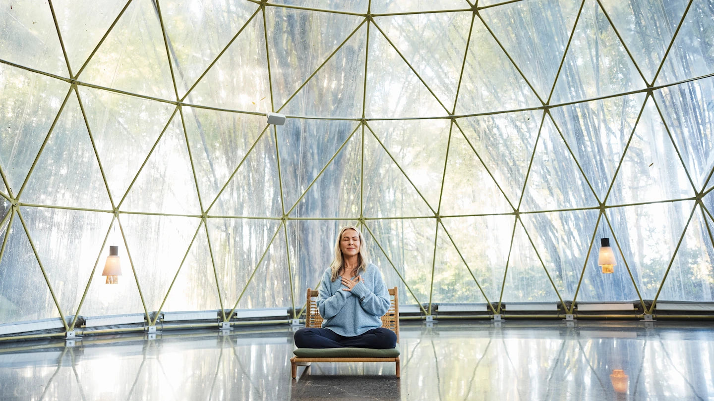 Lorna Jane sits cross-legged on a cushioned wooden chair inside a bright glass-domed space, with her eyes closed and hands resting over her chest in a calm, meditative pose.