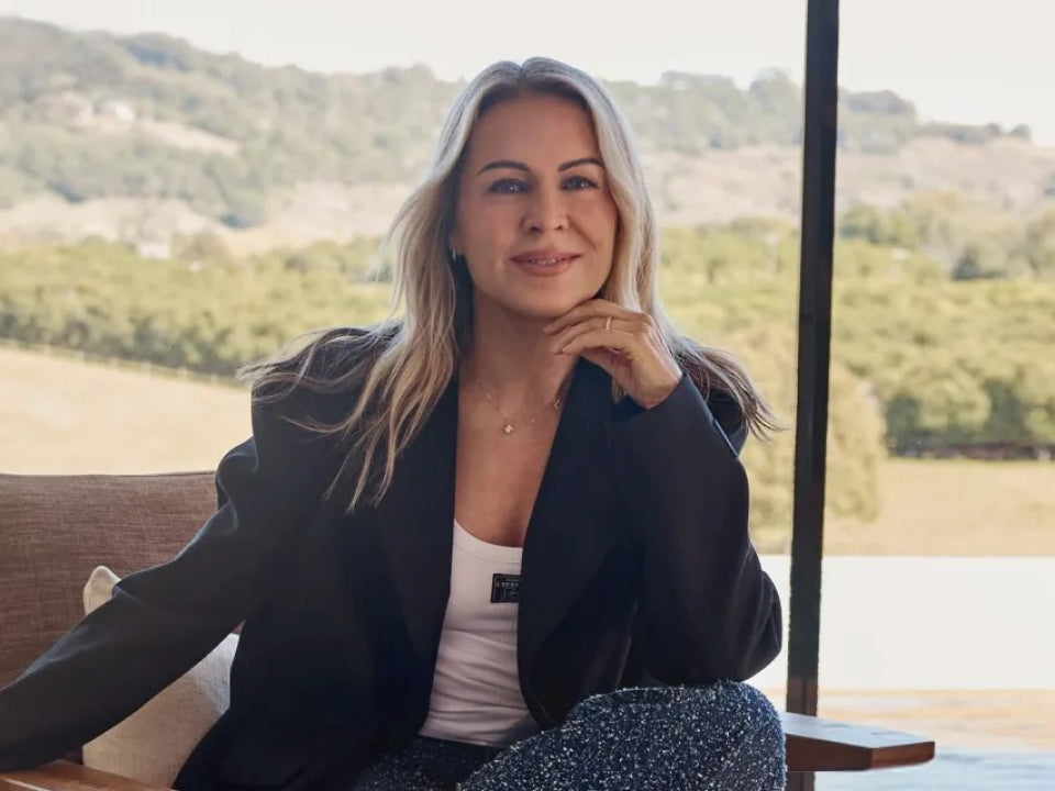 Lorna Jane sits indoors by a large window, smiling with her chin resting on her hand. She wears a black blazer over a white top, with a scenic view of trees and rolling hills in the background.