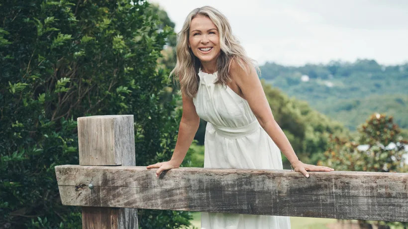 Lorna Jane in a flowing white sleeveless dress leans on a wooden fence outdoors, with lush green trees and rolling hills in the background.