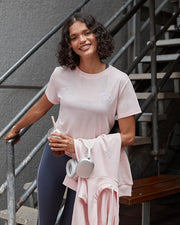 Model wearing a light pink t shirt with blue leggings, holding a light pink jacket and coffee