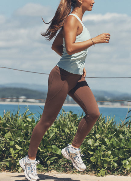 Model wearing a light blue tank with brown leggings, running on a path