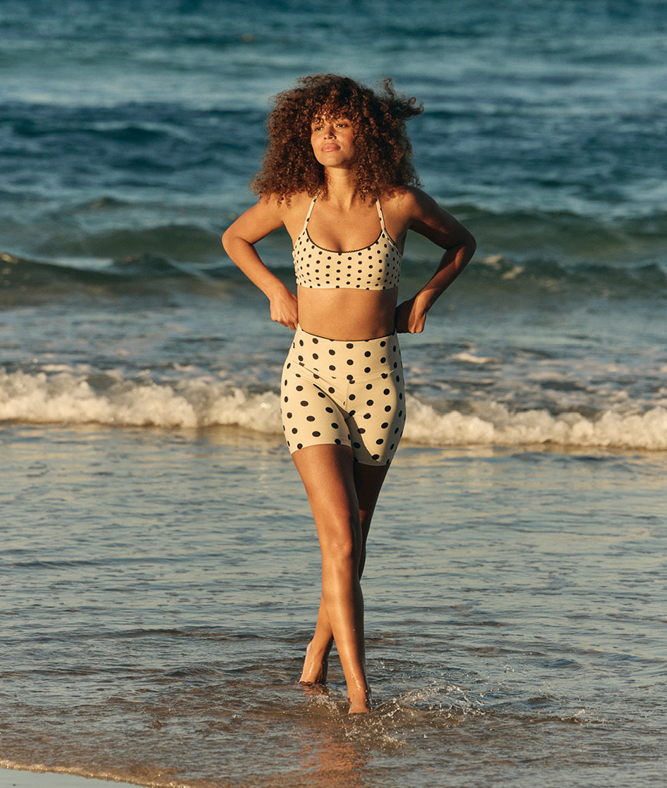 Model wearing a white and black polka dot sports bra and bike shorts, standing in the water at the beach