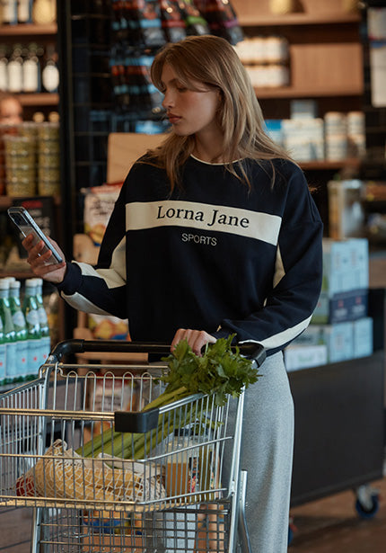 Woman in a navy Lorna Jane Sports sweatshirt pushing a grocery cart of fresh produce while looking at her phone in a store.