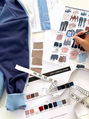 Flat lay of a Lorna Jane design workspace with a blue colour-blocked sweatshirt, fabric swatches, a garment tech sketch sheet, colour palette cards and a tape measure, as a hand sketches with a pencil.