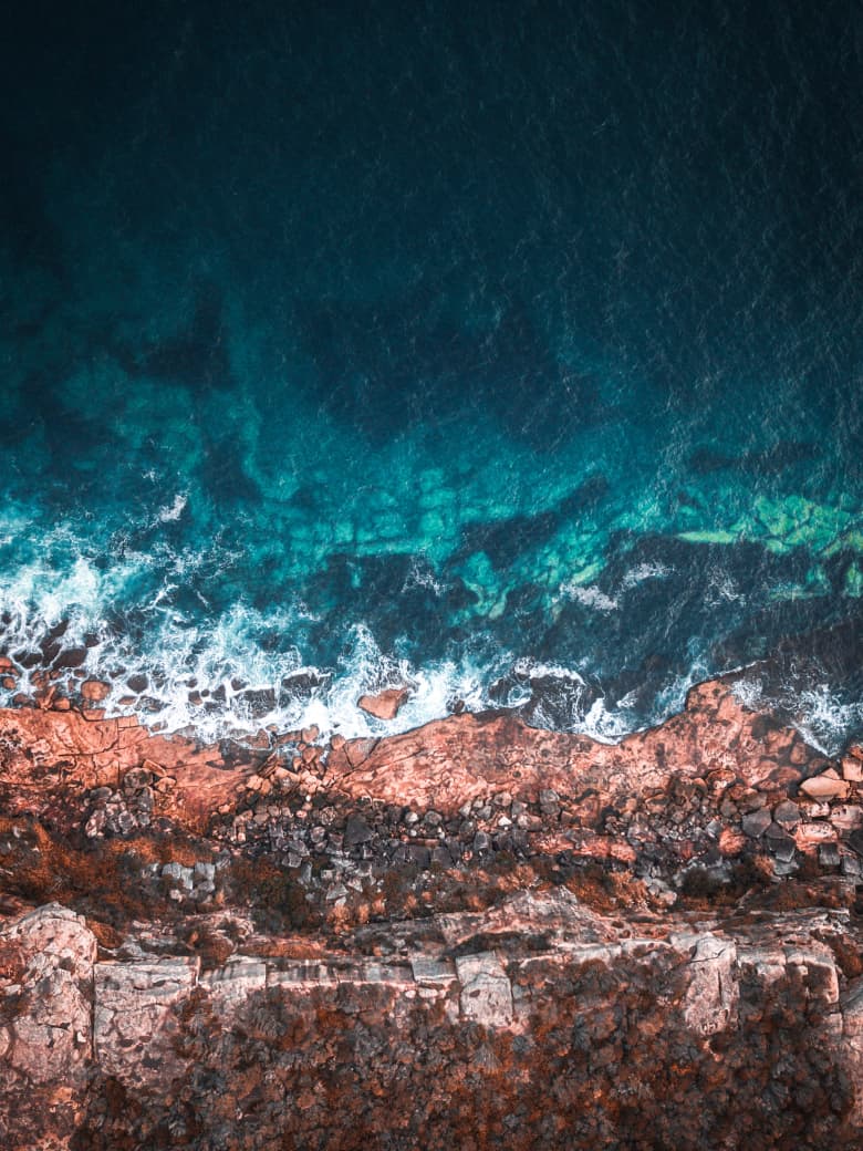 Aerial view of deep teal ocean water meeting a rugged rocky coastline with white breaking waves.