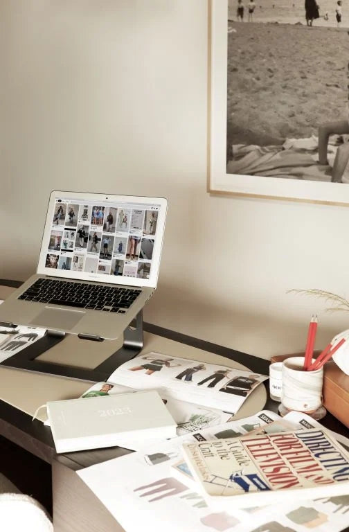 A home desk with a laptop on a stand showing a mood board, alongside book, a magazine, a planner and a pencil cup.