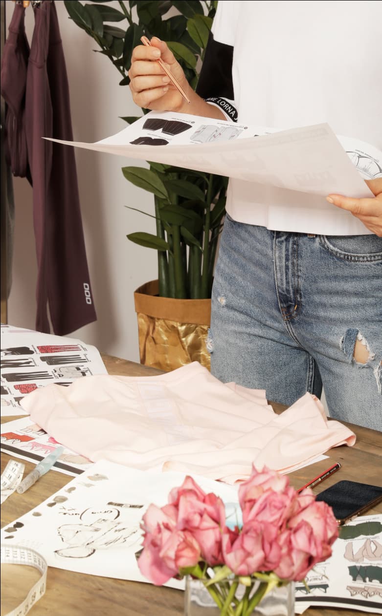 Person reviewing apparel design sketches beside a table with fabric samples, flowers and a folded pink garment.