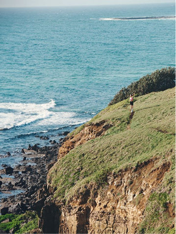 A lone runner on a grassy coastal cliff top overlooking turquoise ocean and a rocky shoreline.