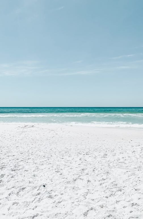 Calm white-sand beach with gentle turquoise waves under a pale blue sky.