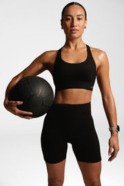 Model wearing a black sports bra and bike shorts, holding an exercise medicine ball