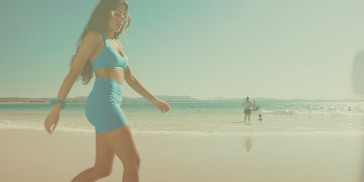 Women in colourful activewear at a sunny beach, walking along the shoreline, carrying a surfboard and playing beach volleyball.