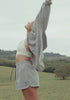 A woman moves through a rolling green valley and lush tropical garden in three elevated warm weather looks, a white crop top with gingham shorts and an oversized striped shirt, a white silk slip dress, and a textured white knit top with striped shorts. Lorna Jane lifestyle activewear. Text: Just Landed: New Season. Shop New Arrivals.