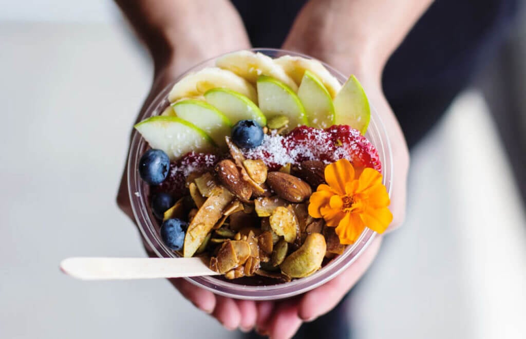 hands holding a chia pudding topped in seasonal fruit, coconuts, nuts and a spoon
