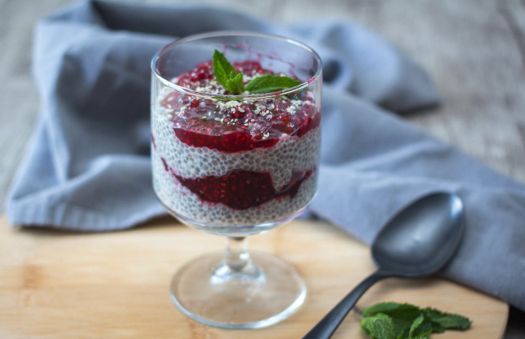 chia seed pudding in stemmed glass layered with strawberry jam on chopping board with spoon